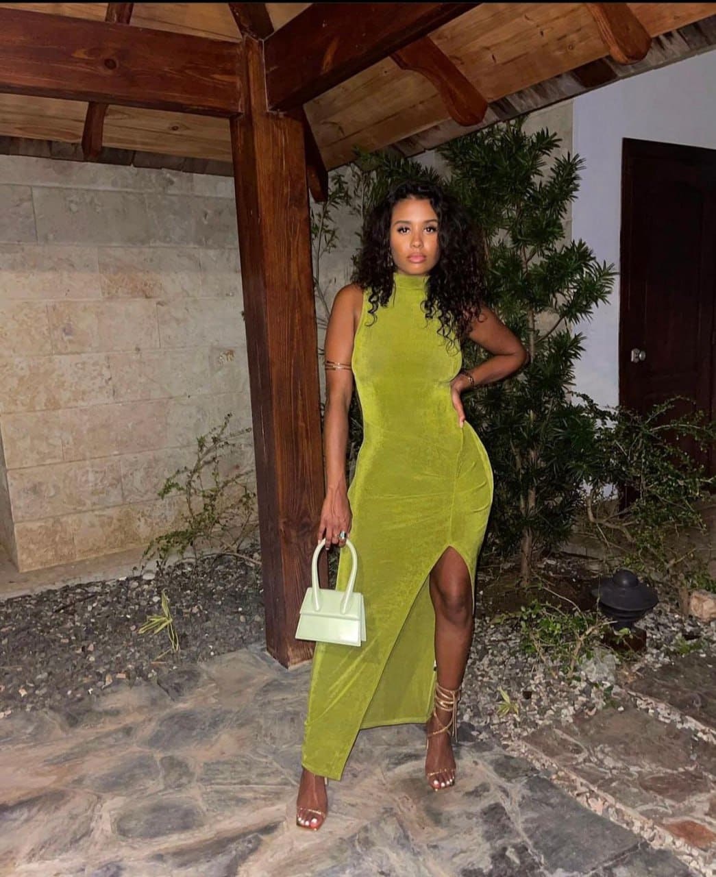 Penelope stands on a stone patio under a pergola, wearing a lime green velvet dress with a slit and holding a light green handbag.
