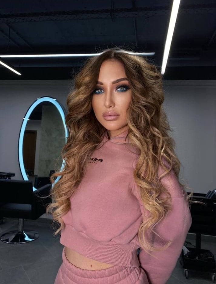 A blonde woman in a pink cropped sweatshirt and pants stands in a salon, with an illuminated mirror and styling chairs behind her.