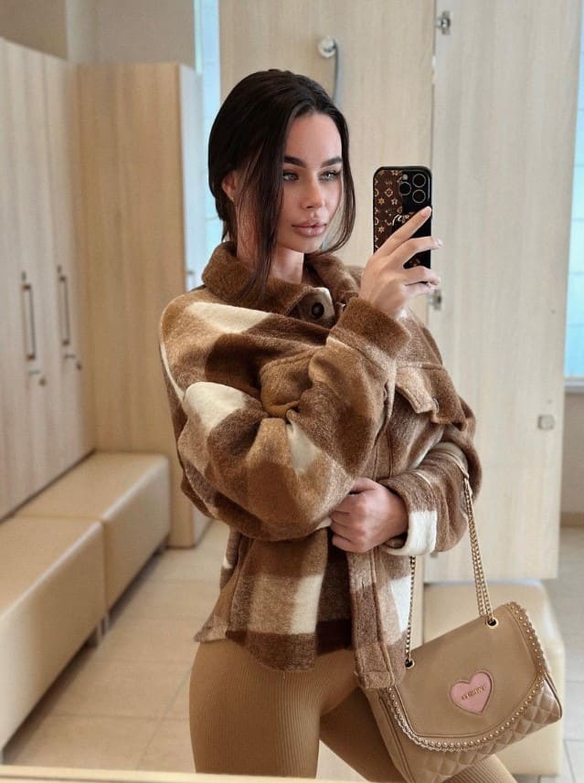 A woman takes a mirror selfie in a plaid fleece jacket and tan leggings, holding a beige quilted handbag in a room with wood cabinets.