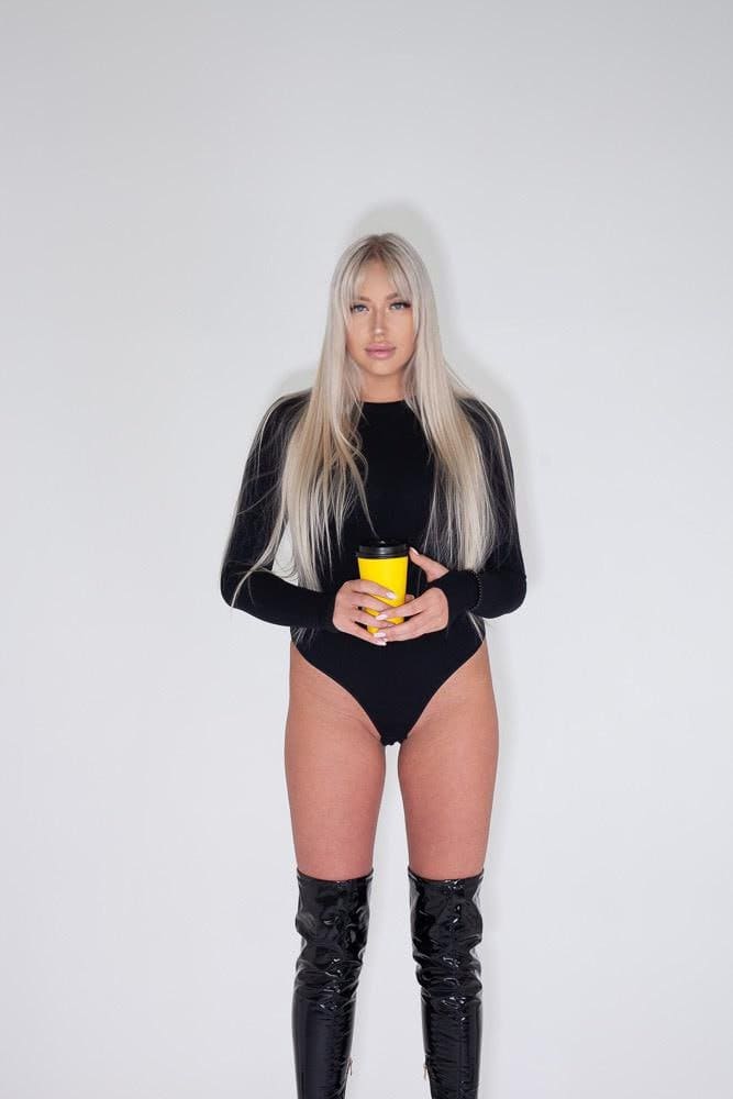 A woman with platinum-blonde hair stands in a black bodysuit and thigh-high boots, holding a yellow travel mug.