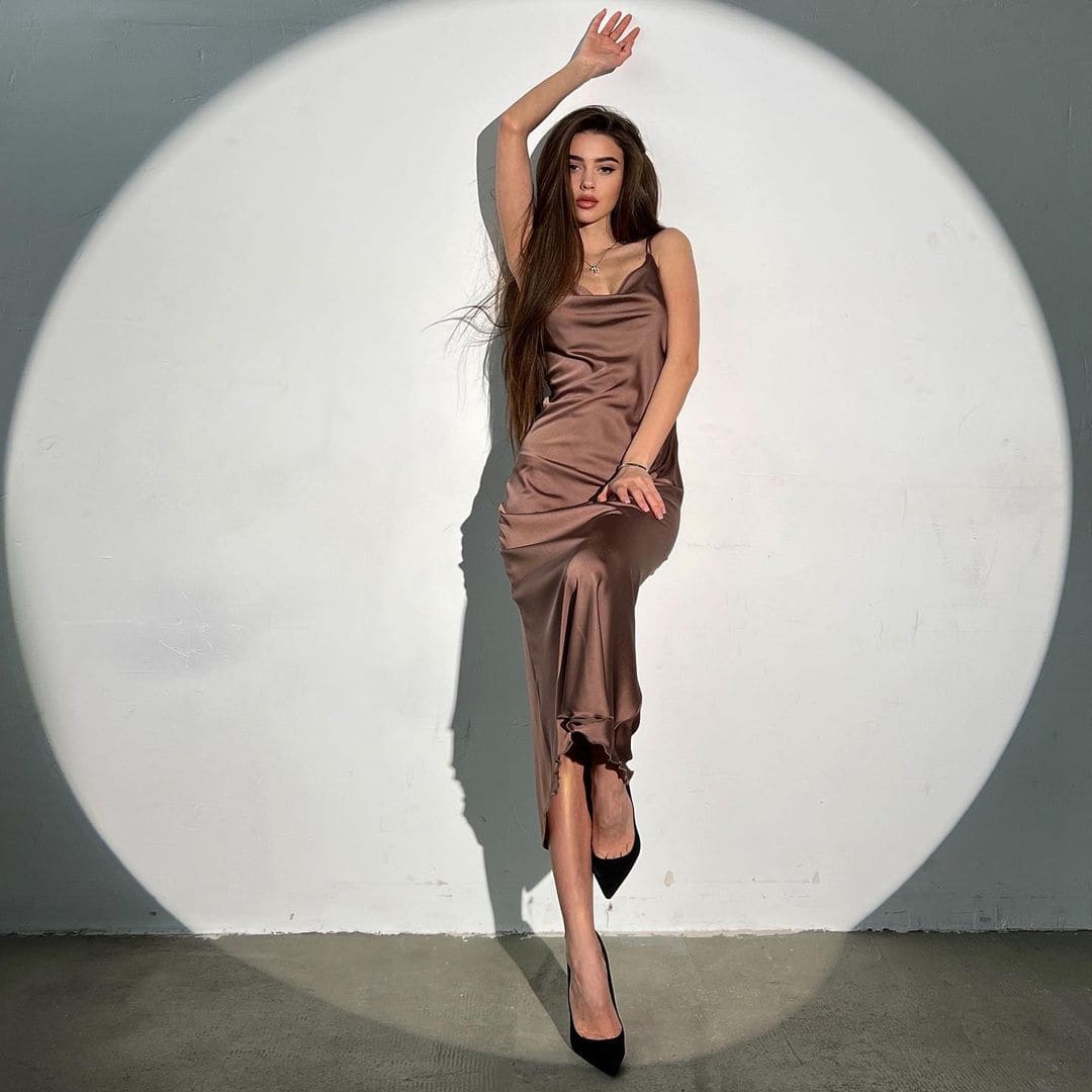 Woman in a brown satin slip dress with a slit stands in a spotlight against a white wall.