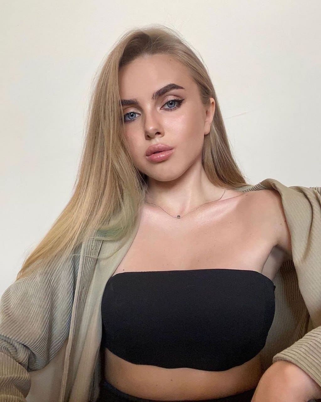 A blonde woman in a black tube top and beige cardigan poses against a plain light wall, wearing a necklace.