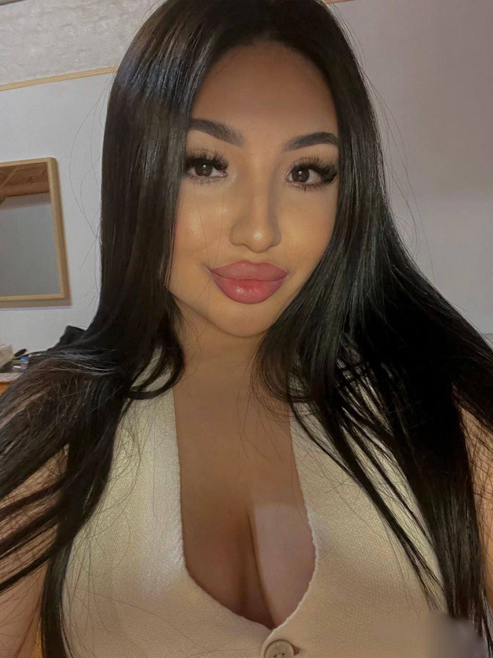 A close-up selfie of a woman in a cream V-neck top, taken in a room with a mirror on the wall behind her.