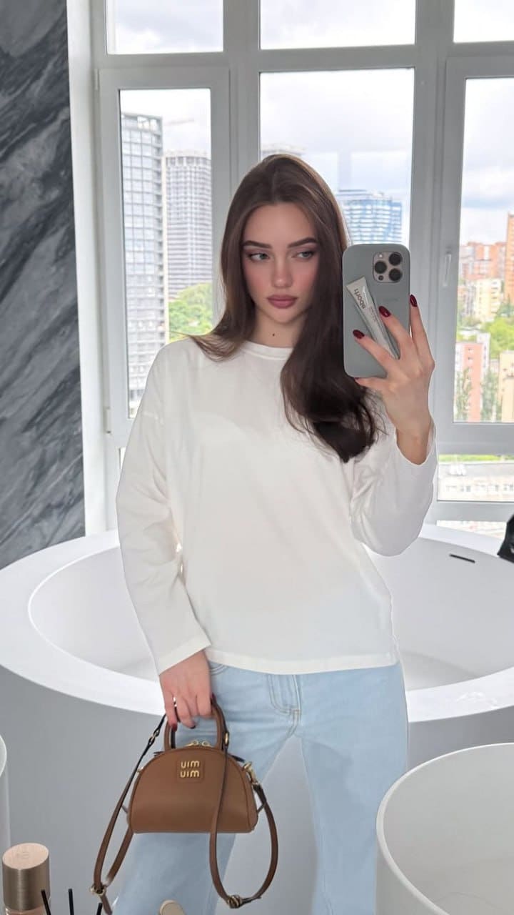 Chloe stands in a bathroom with a bathtub and window, taking a mirror selfie in a white long-sleeved top, light blue jeans, and holding a brown handbag.
