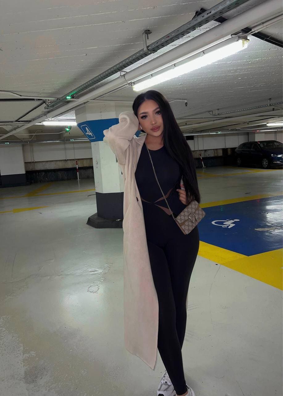 A woman in a black bodysuit, pink coat, and sneakers stands in a parking garage holding a crossbody bag.