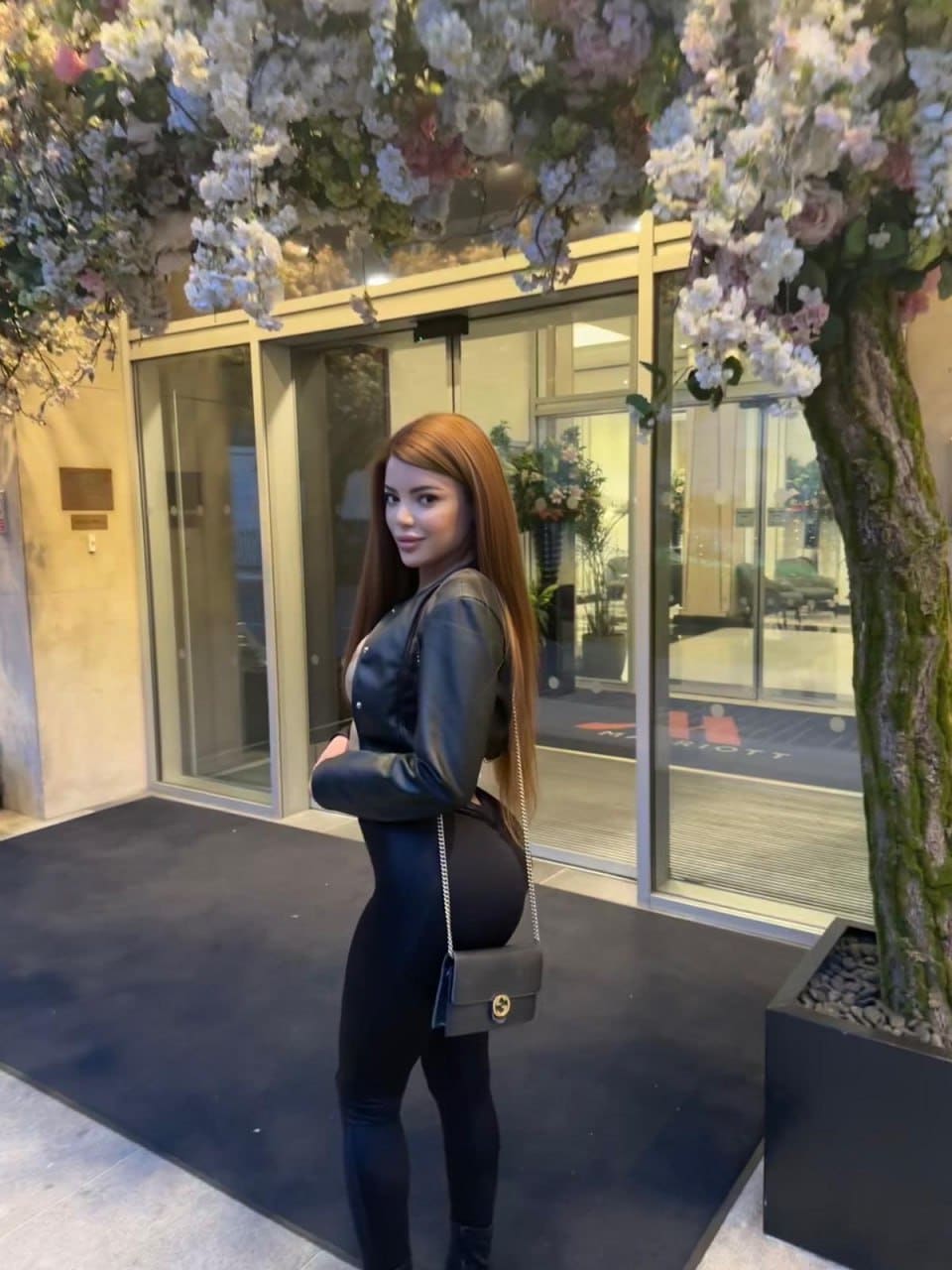A woman in profile wears a black leather top and leggings with a chain bag, standing before a glass entrance with flowers.
