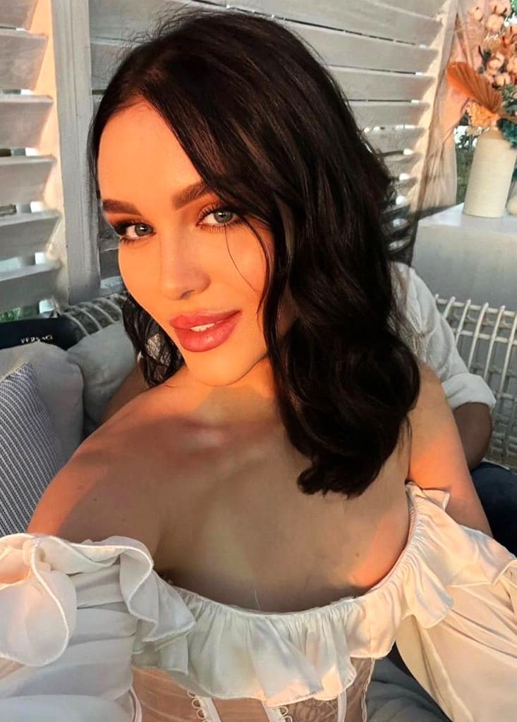 A woman in a white off-the-shoulder ruffled top sits indoors near a window with blinds and a vase of flowers.