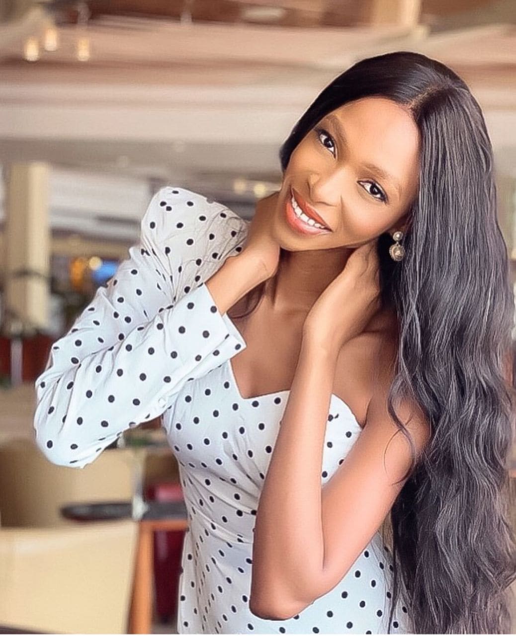 Candace smiles indoors in a white polka-dot dress with one puffed sleeve and one bare shoulder, hands near her neck.