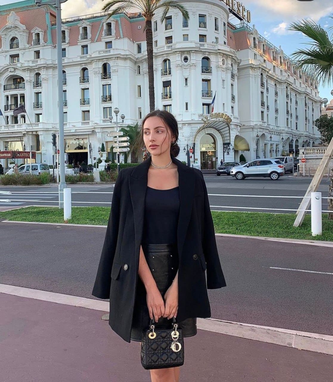 A woman stands on a city street in a black blazer and skirt, holding a handbag.