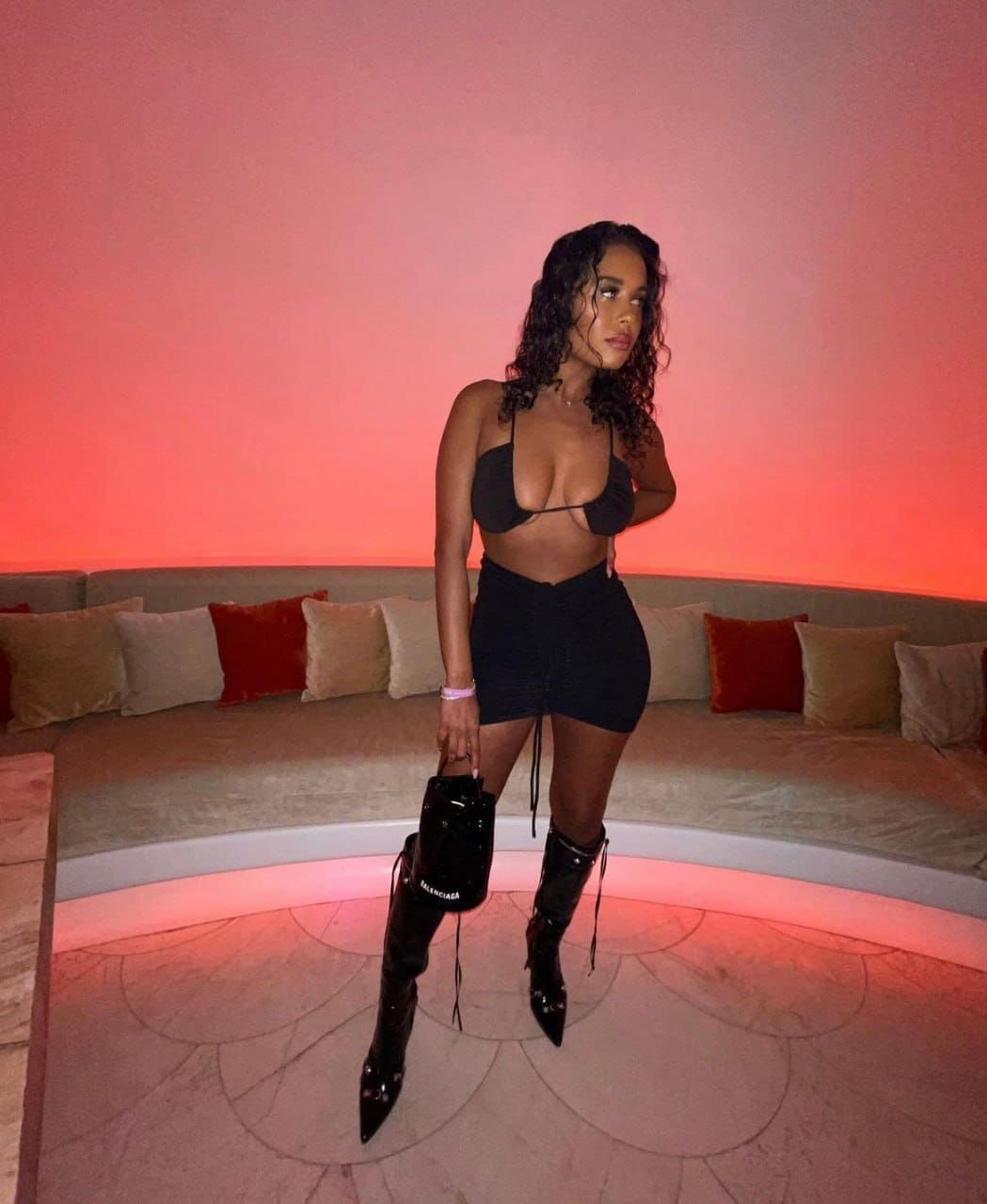 A woman in a black bra top, short skirt, and lace-up boots stands in a lounge with pink-lit walls, holding a small black handbag.