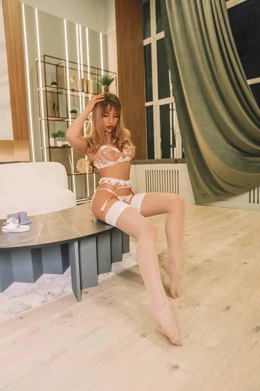 Limma sits on a low table in a modern room, wearing white lace lingerie and thigh-high stockings, with one hand raised to her head.