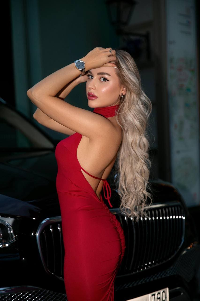 A blonde woman in a backless red dress stands before a black car, raising her right hand to her forehead.