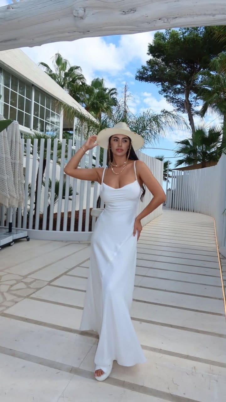 A woman in a long white dress and straw hat stands on a tiled walkway, touching her hat brim against palm trees.