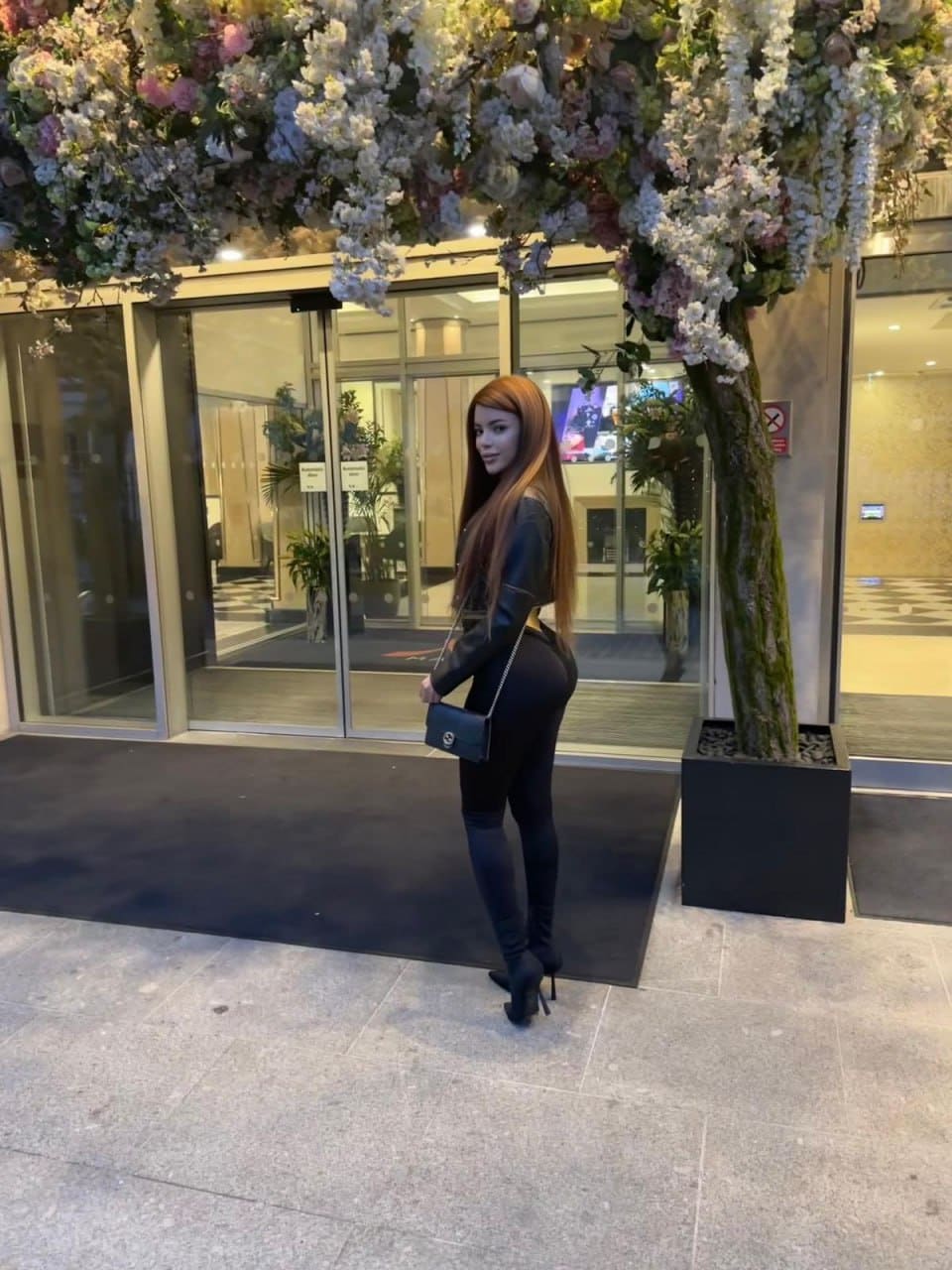 A woman in a black leather jacket, leggings, and high-heeled boots stands on a walkway near a floral arrangement and tree.