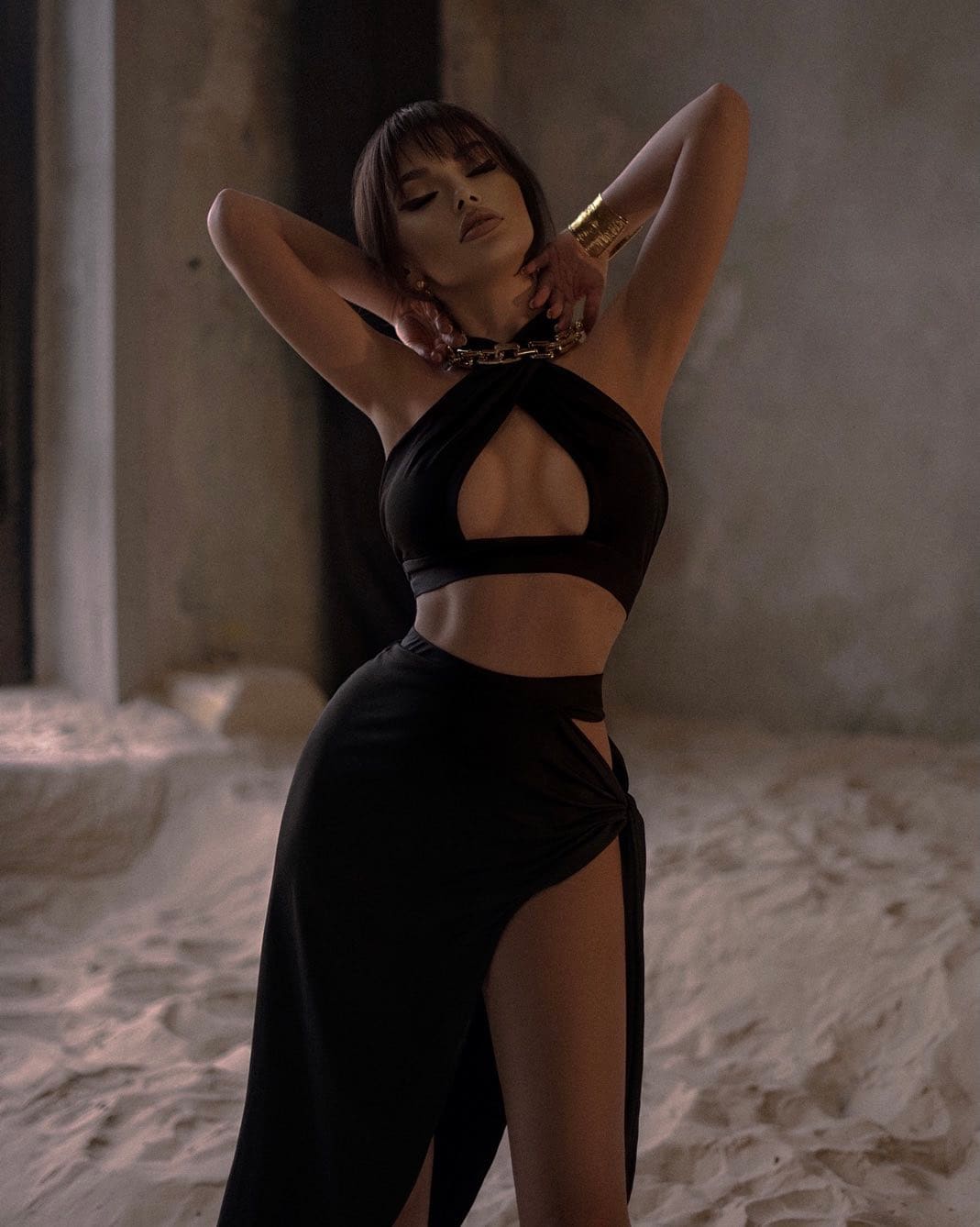 A woman with bangs stands in a dim concrete room wearing a black cut-out halter top and high-slit skirt, with arms raised behind her head.