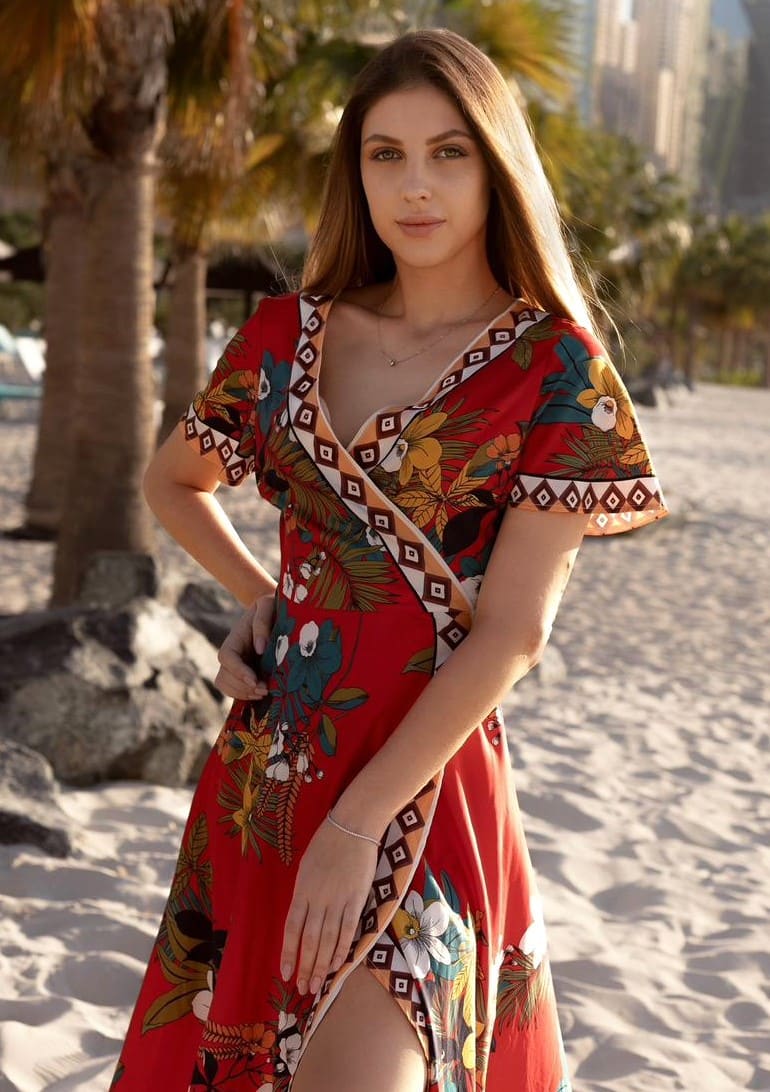 Marika stands on a sandy beach in a red floral wrap dress with a high slit, hand on hip, framed by palm trees and distant buildings.