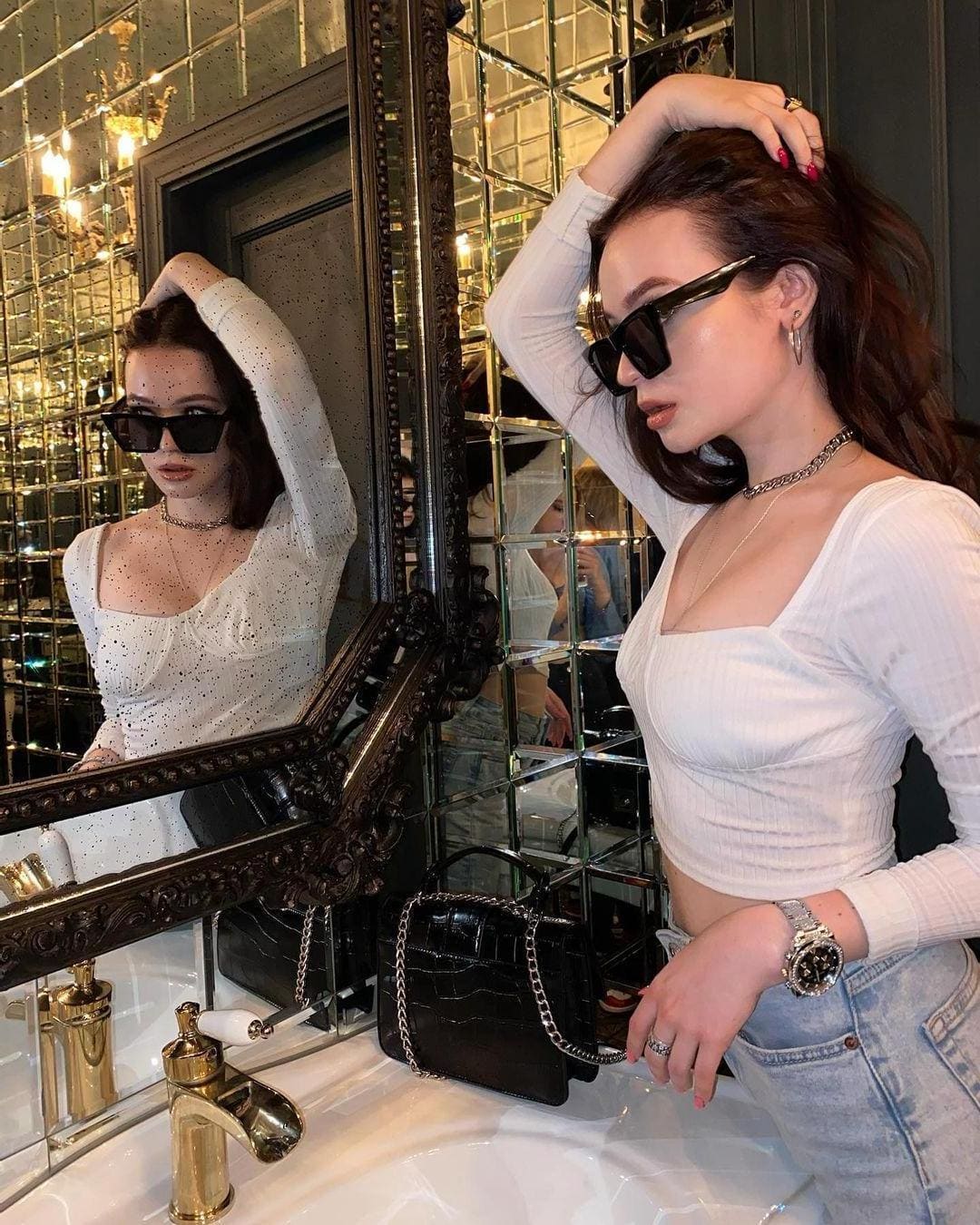 A woman in a white crop top and jeans stands before an ornate mirror in a bathroom, wearing sunglasses and touching her hair.