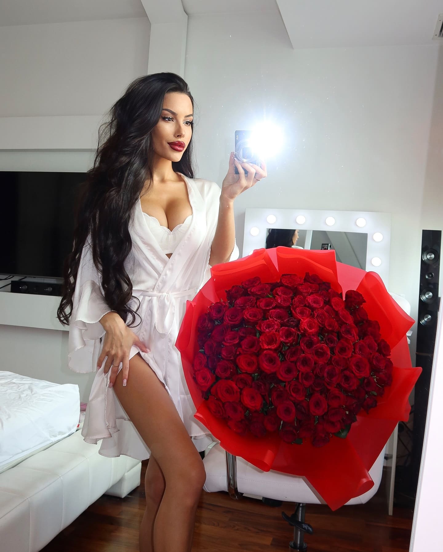 In a bedroom, she holds a phone in a short white robe, with a large bouquet of red roses on a chair nearby.